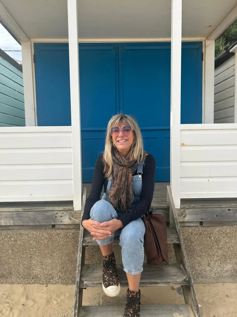 Joanne smiling on the steps of a blue beach hut, in denim dungarees and a paisley scarf.