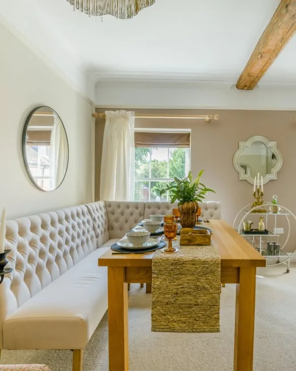 The dining room at Willow: a long tufted banquette, an oak table laid for guests, a sash window onto green trees and soft blush walls.